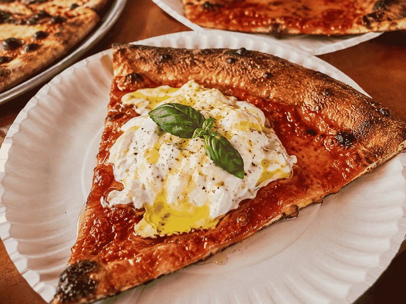 L’Industrie Pizzeria burrata pizza slice with tomato sauce, fresh basil, olive oil, and crispy charred crust.